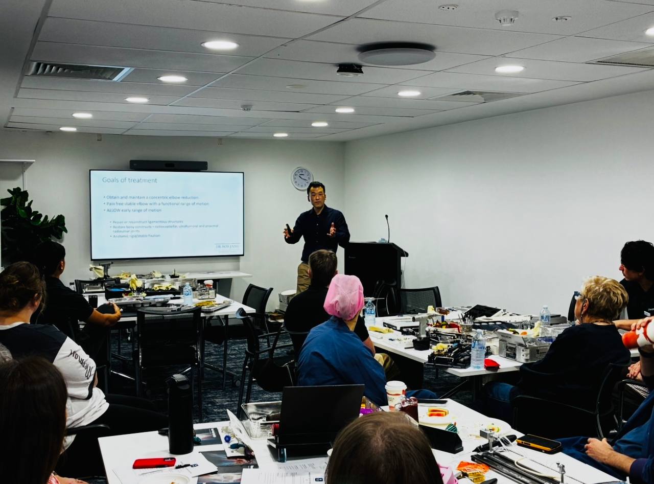 Dr Bob Jang presenting to theatre nursing staff at the Orthopaedic Theatre Education Day, with a slide on the goals of treatment for elbow injuries behind him.