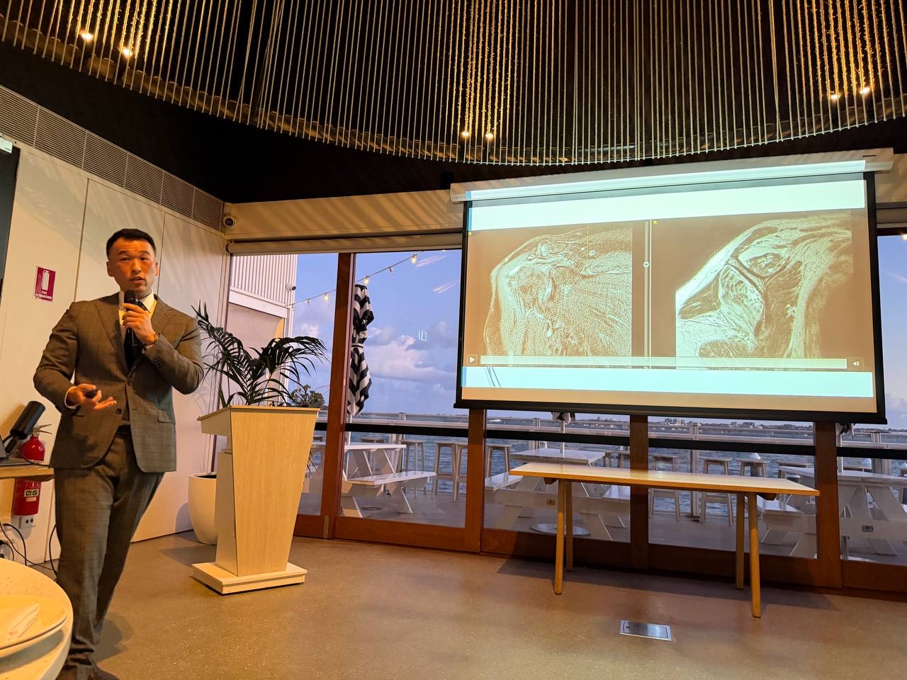 Dr Bob Jang presenting at an educational evening, with shoulder MRI scans on the screen beside him.